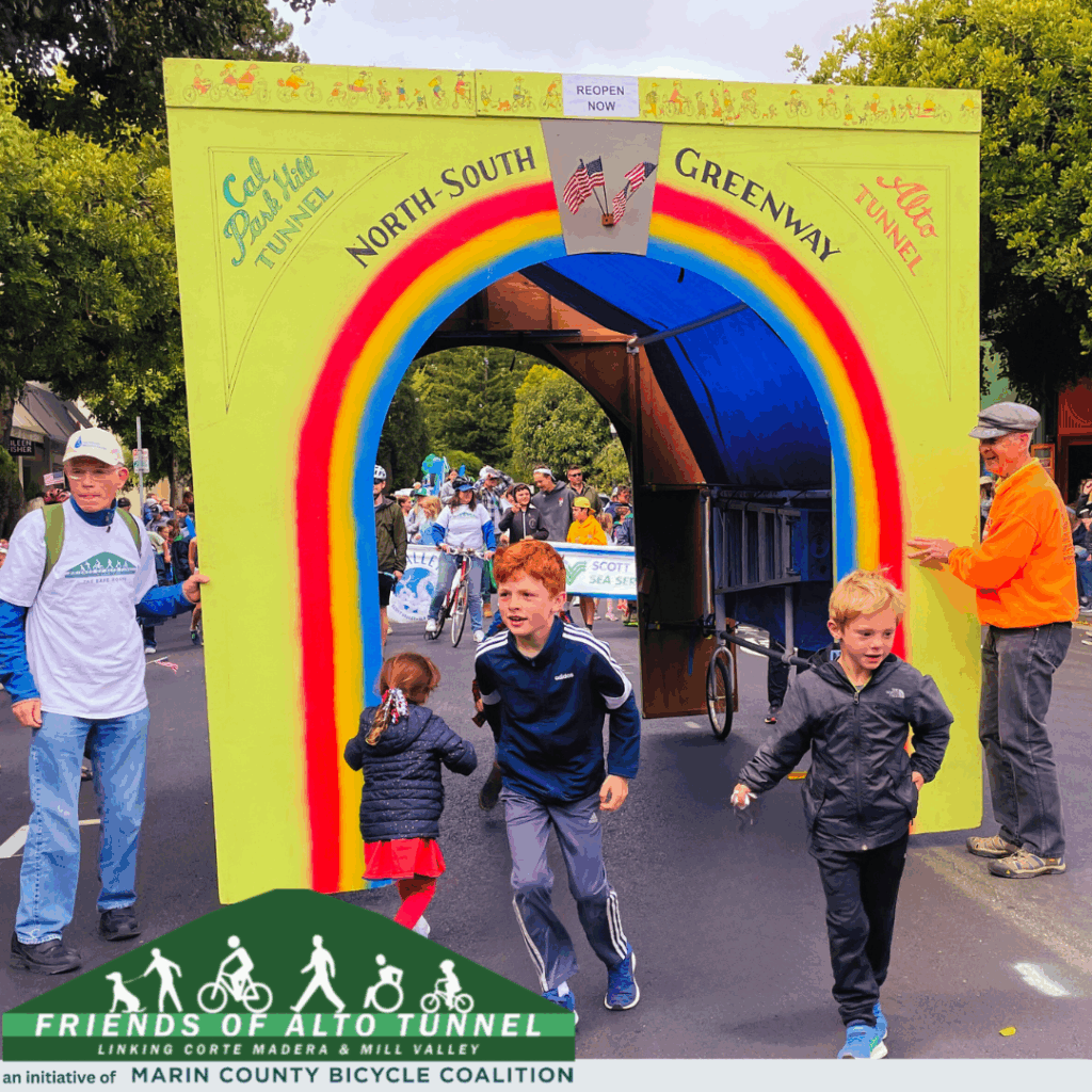 Community event with families and cyclists passing through rainbow-themed Alto Tunnel display supporting North-South Greenway.