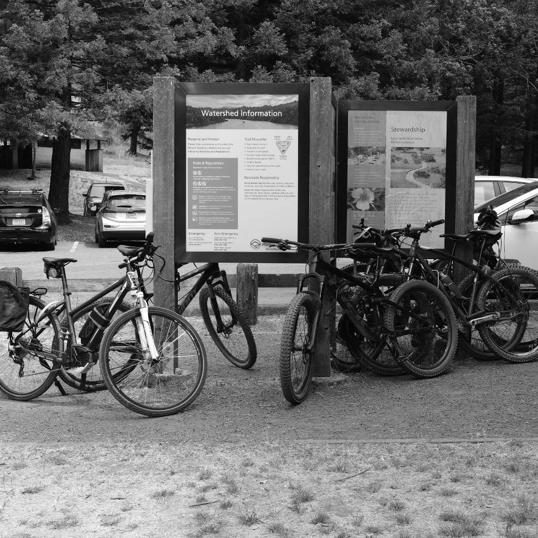 Bicycles parked near an information board at the Mt. Tam Watershed, with signs about trail rules and stewardship, and cars visible in the background.