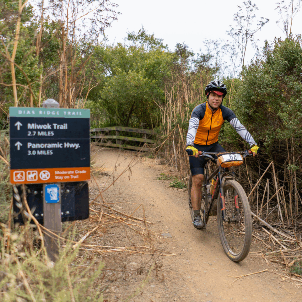 Cyclist riding past trailhead sign marking Dias Ridge Trail with connections to Miwok Trail and Panoramic Highway